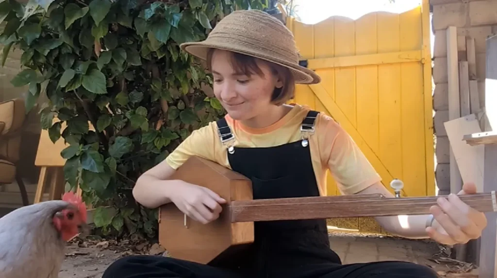 Phoebe’s music video, fretless banjo, charming chicken, Down in the River to Pray, authenticity, whimsical, homely vibe, heartfelt performance, simplicity, nostalgia.